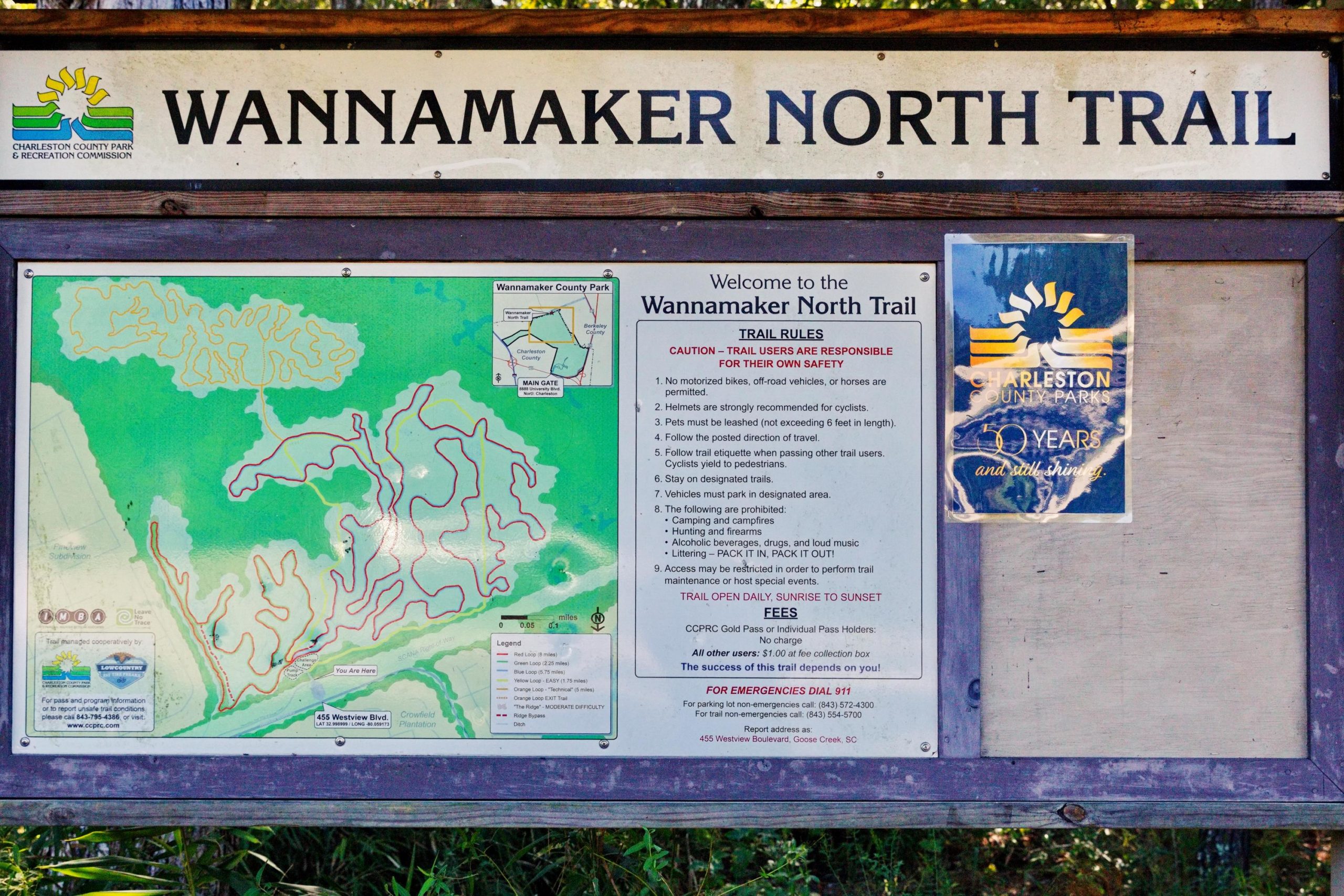 A signboard for the Wannamaker North Trail, featuring a map of the trail and guidelines for users. The sign includes trail rules such as safety precautions for cyclists and information on permitted activities. It displays the Charleston County Park & Recreation Commission logo and details on fees and emergency contact information. The background shows lush greenery typical of a park setting. Wannamaker Park mountain bike trail.