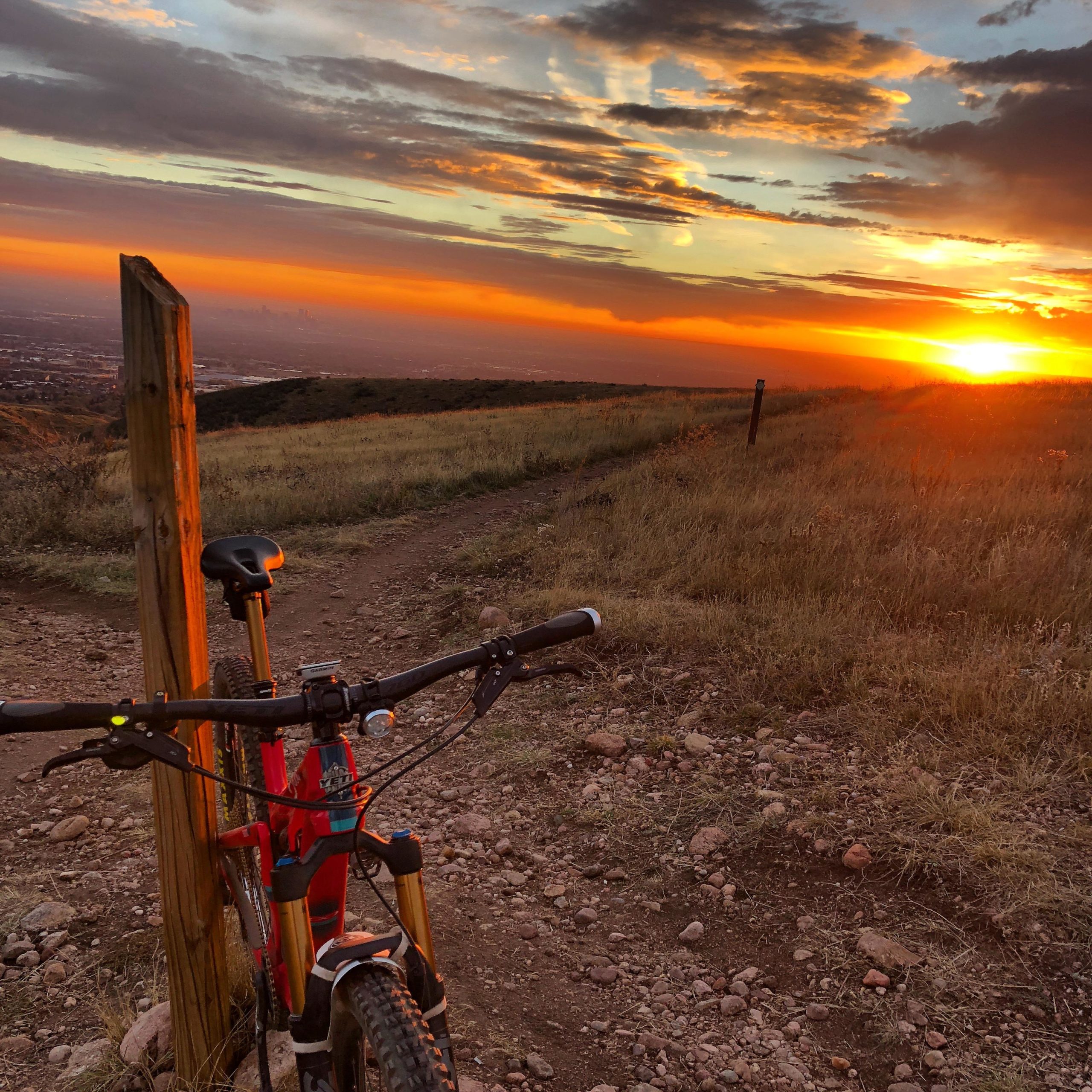 A mountain bike rests against a wooden post on a rugged dirt path, overlooking a vast landscape bathed in the warm colors of a sunset. The sky is painted with hues of orange, pink, and purple as the sun dips below the horizon. Tall grasses frame the trail, and a distant city skyline can be faintly seen in the background. Green Mountain mountain bike trail.