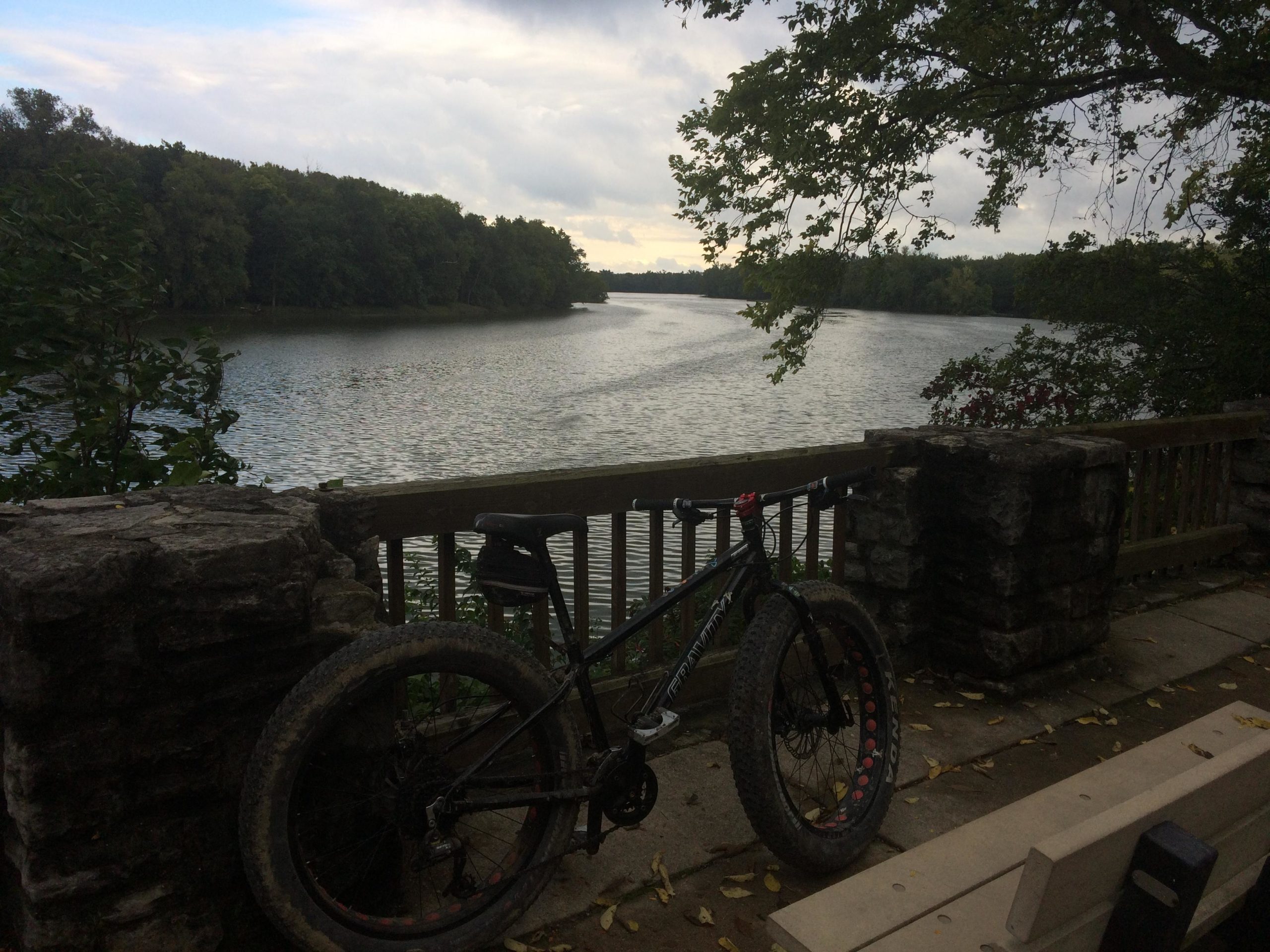 A fat bike resting against a stone railing overlooking a calm river, surrounded by greenery, with a cloudy sky in the background. The scene captures a peaceful outdoor setting, ideal for cycling and nature appreciation. Farnsworth mountain bike trail.