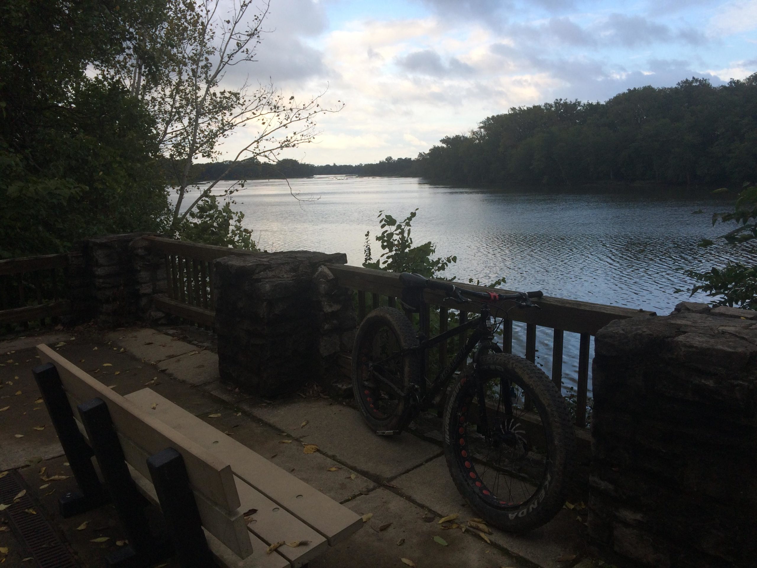 A serene lakeside view featuring a wooden bench and a mountain bike resting against a stone railing. The calm water reflects the cloudy sky and surrounding trees, creating a peaceful outdoor atmosphere. Farnsworth mountain bike trail.