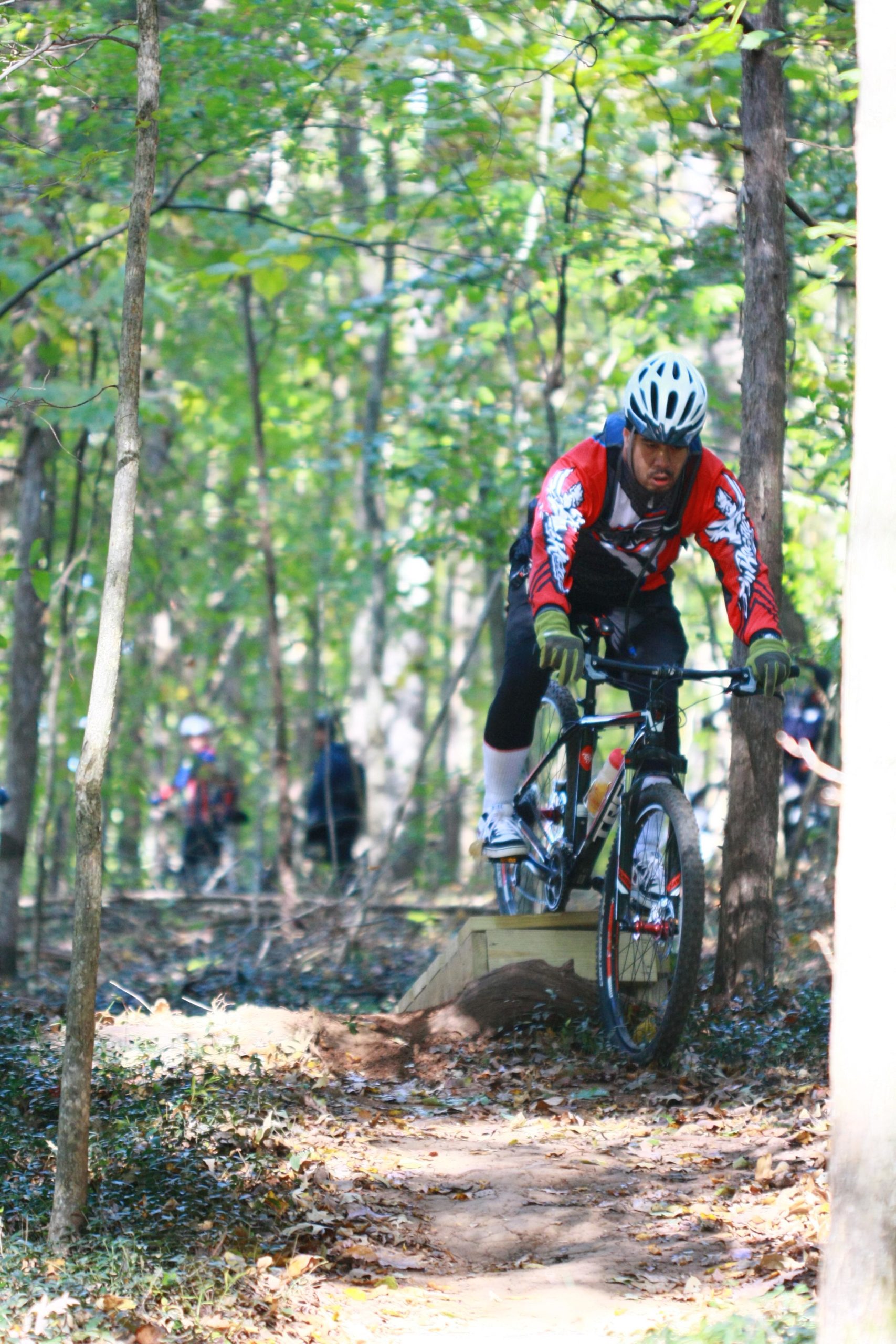 A mountain biker in a red jersey and helmet is skillfully navigating a trail in a wooded area, riding over a small wooden ramp. Surrounding trees provide natural shade, while other cyclists can be seen in the background, enjoying the trail. The scene captures the excitement and challenge of mountain biking in nature. Tanglewood Park mountain bike trail.