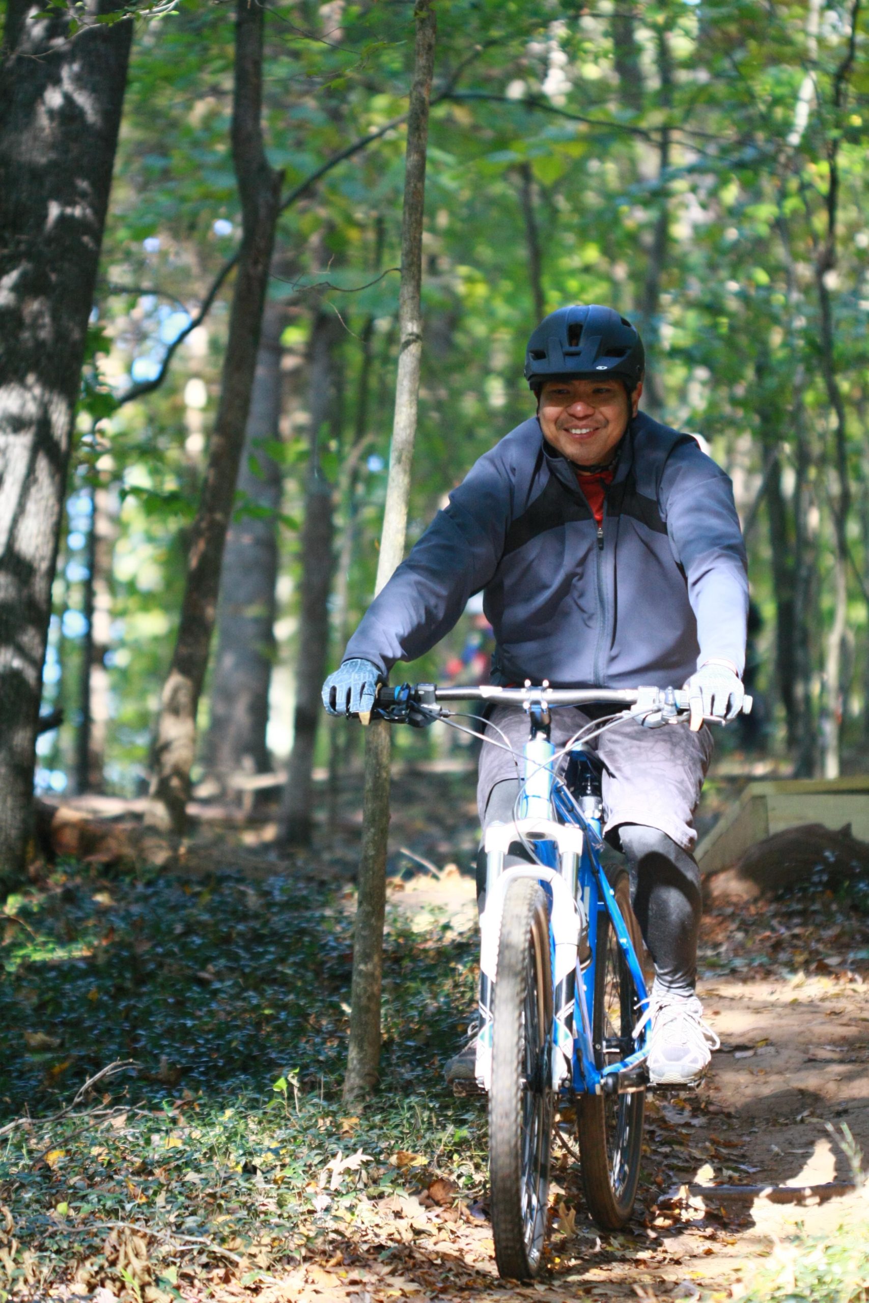 A person riding a blue mountain bike along a dirt trail in a wooded area, smiling while wearing a black helmet and gloves. The surrounding environment features green trees and fallen leaves, indicating a sunny day. Tanglewood Park mountain bike trail.