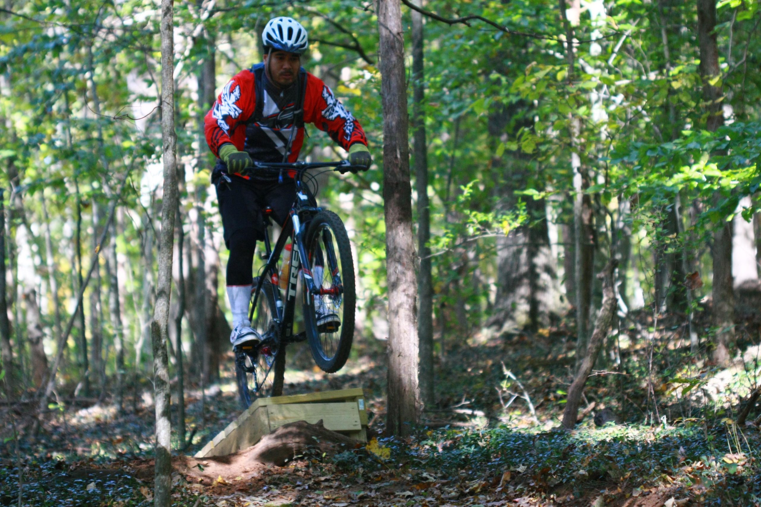 A mountain biker in a red jersey and black shorts is seen jumping off a small wooden ramp in a forested area, surrounded by trees and greenery. The rider is focused and dressed in protective gear, showcasing a dynamic moment of off-road biking. Tanglewood Park mountain bike trail.