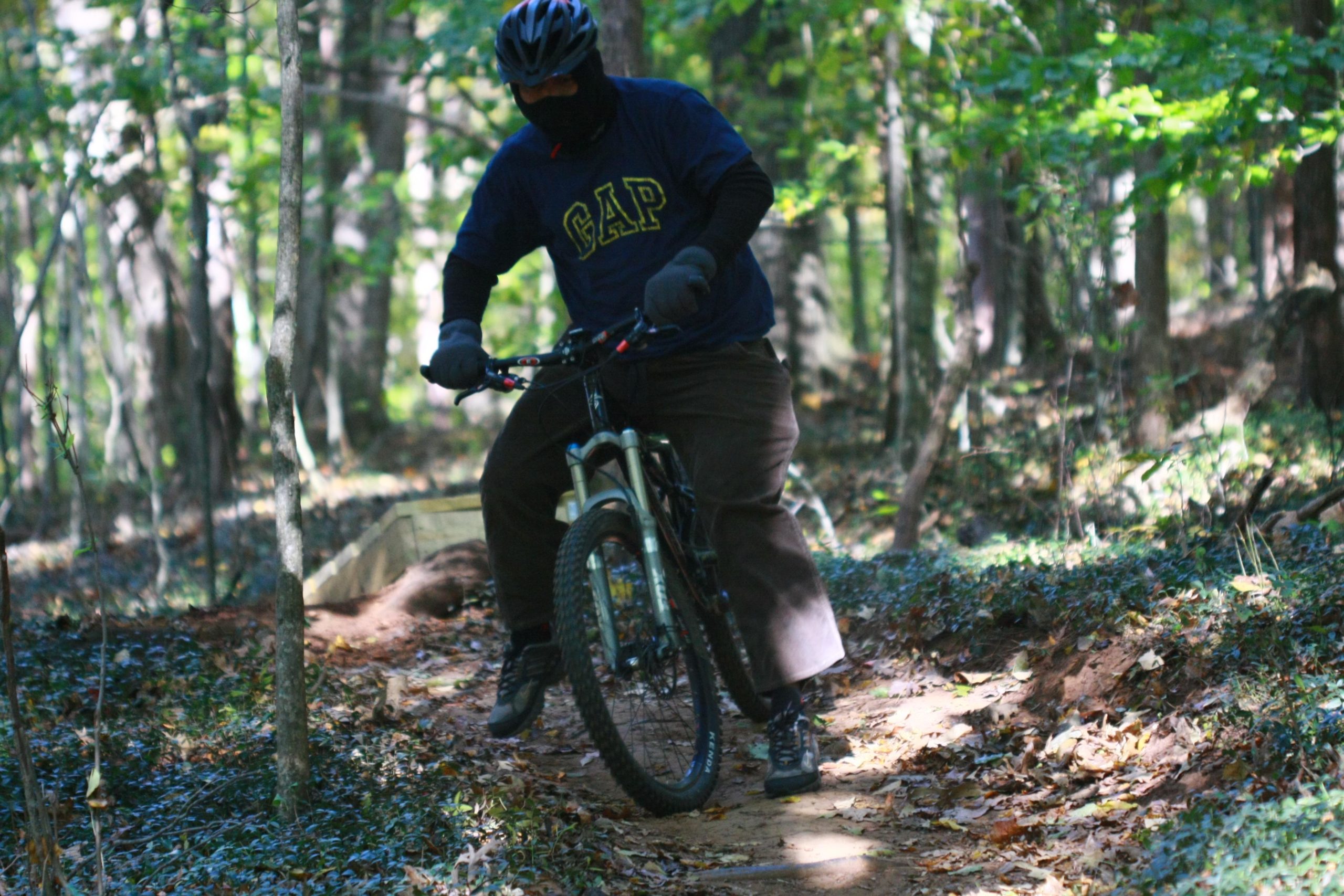 A person wearing a helmet and a face covering rides a mountain bike along a dirt trail in a forested area. Sunlight filters through the trees, illuminating the path and surrounding foliage. Tanglewood Park mountain bike trail.