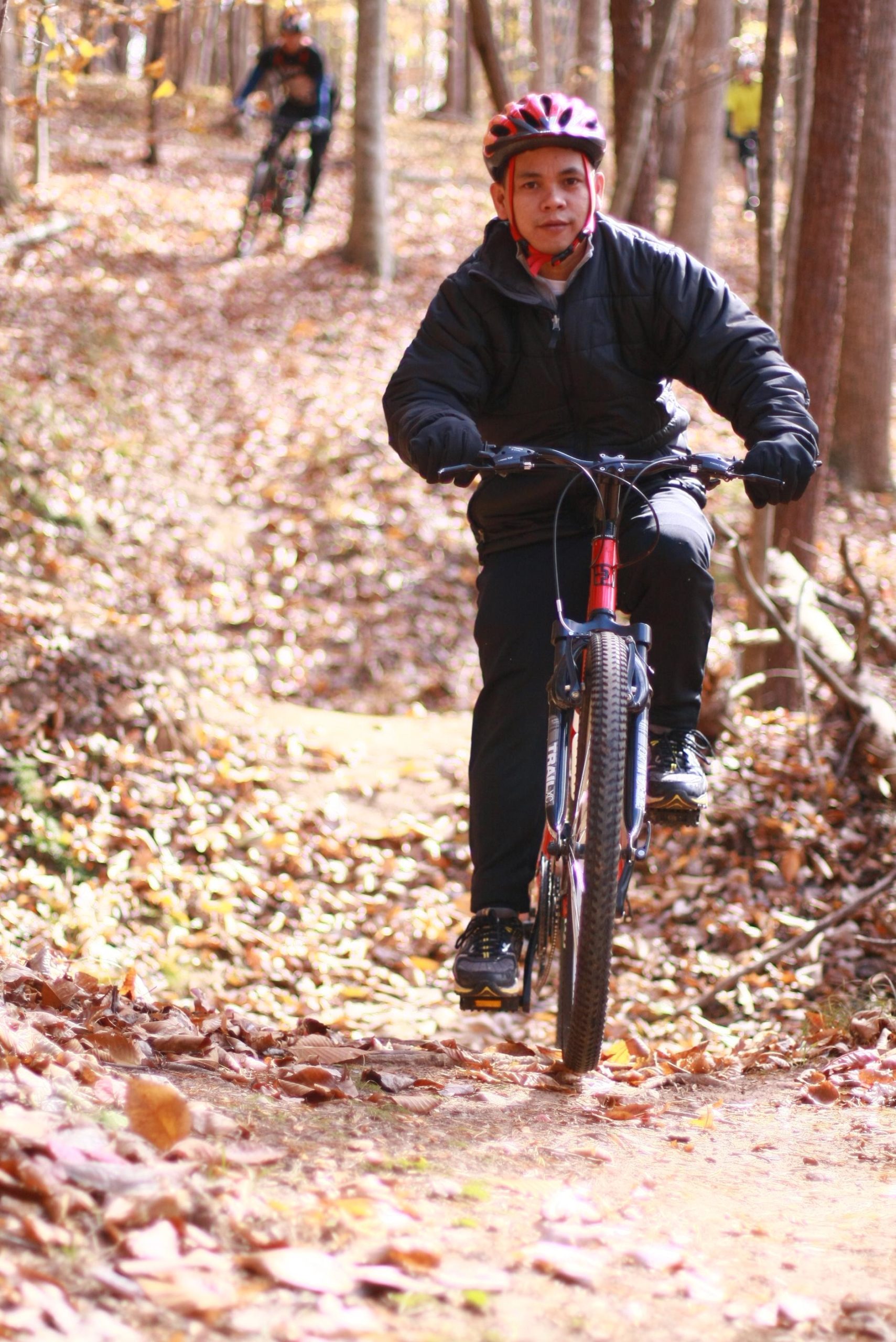 A person riding a mountain bike along a dirt path covered in autumn leaves, with trees in the background. Another cyclist can be seen in the distance. The rider is wearing a helmet and a dark jacket, showcasing an outdoor biking activity in a natural setting. Cedar Ridge Chatmoss mountain bike trail.