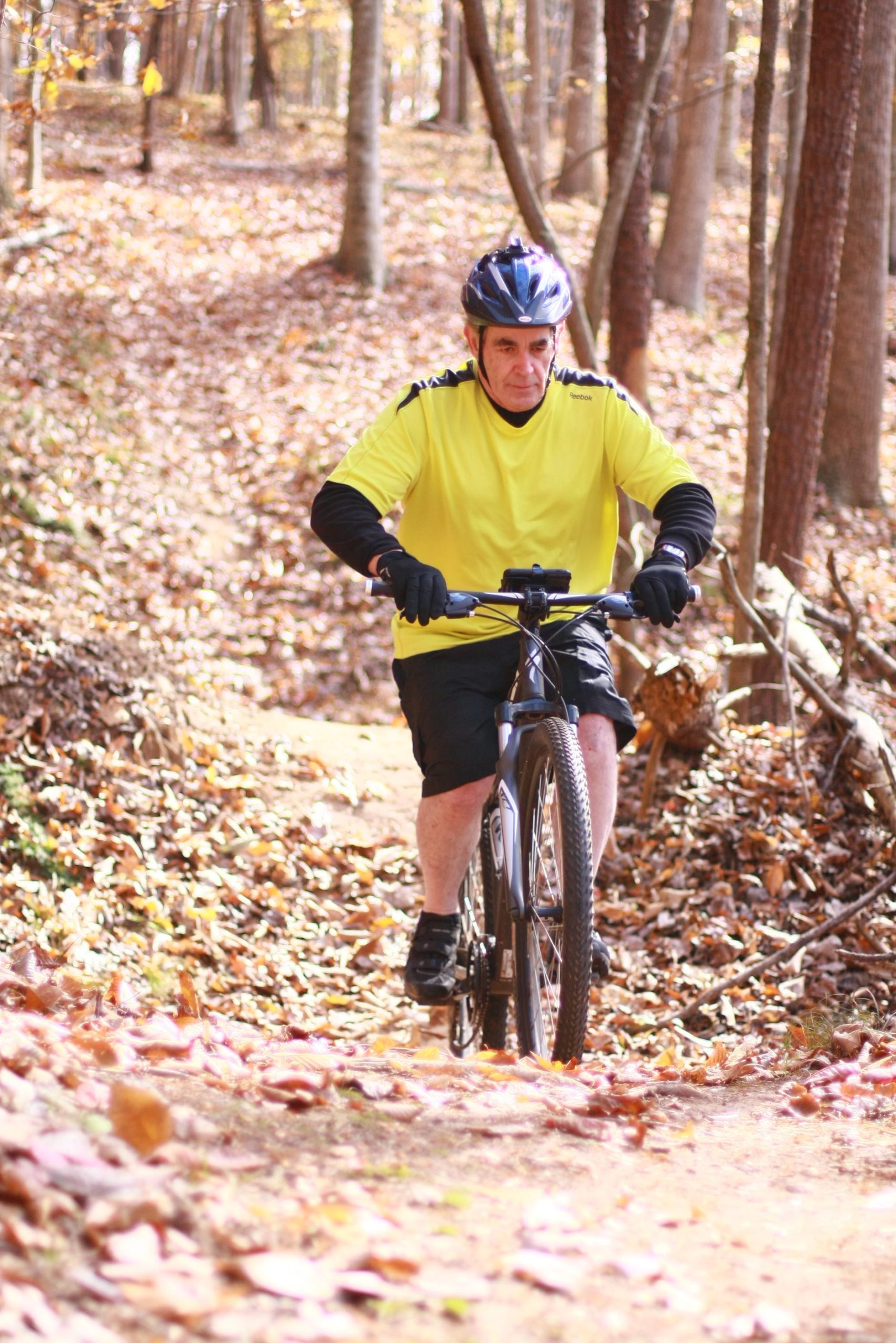An older man in a bright yellow shirt and black shorts rides a mountain bike on a leaf-covered trail in a forest during autumn. He wears a cycling helmet and gloves as he navigates the path surrounded by trees. Cedar Ridge Chatmoss mountain bike trail.