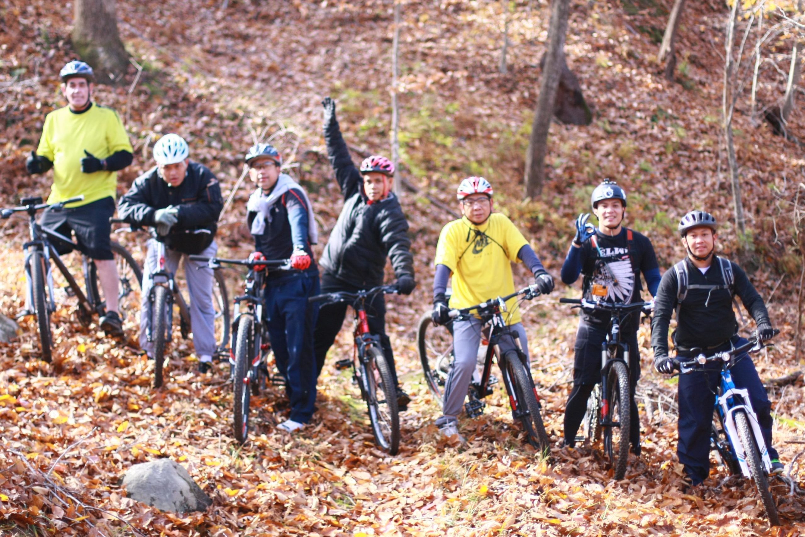 A group of seven cyclists, wearing helmets and casual athletic clothing, pose on a trail covered with autumn leaves. They are gathered with their mountain bikes, showcasing a friendly and energetic atmosphere. Some of them are giving thumbs up or waving, indicating enthusiasm for their biking activity in a forested setting. Cedar Ridge Chatmoss mountain bike trail.