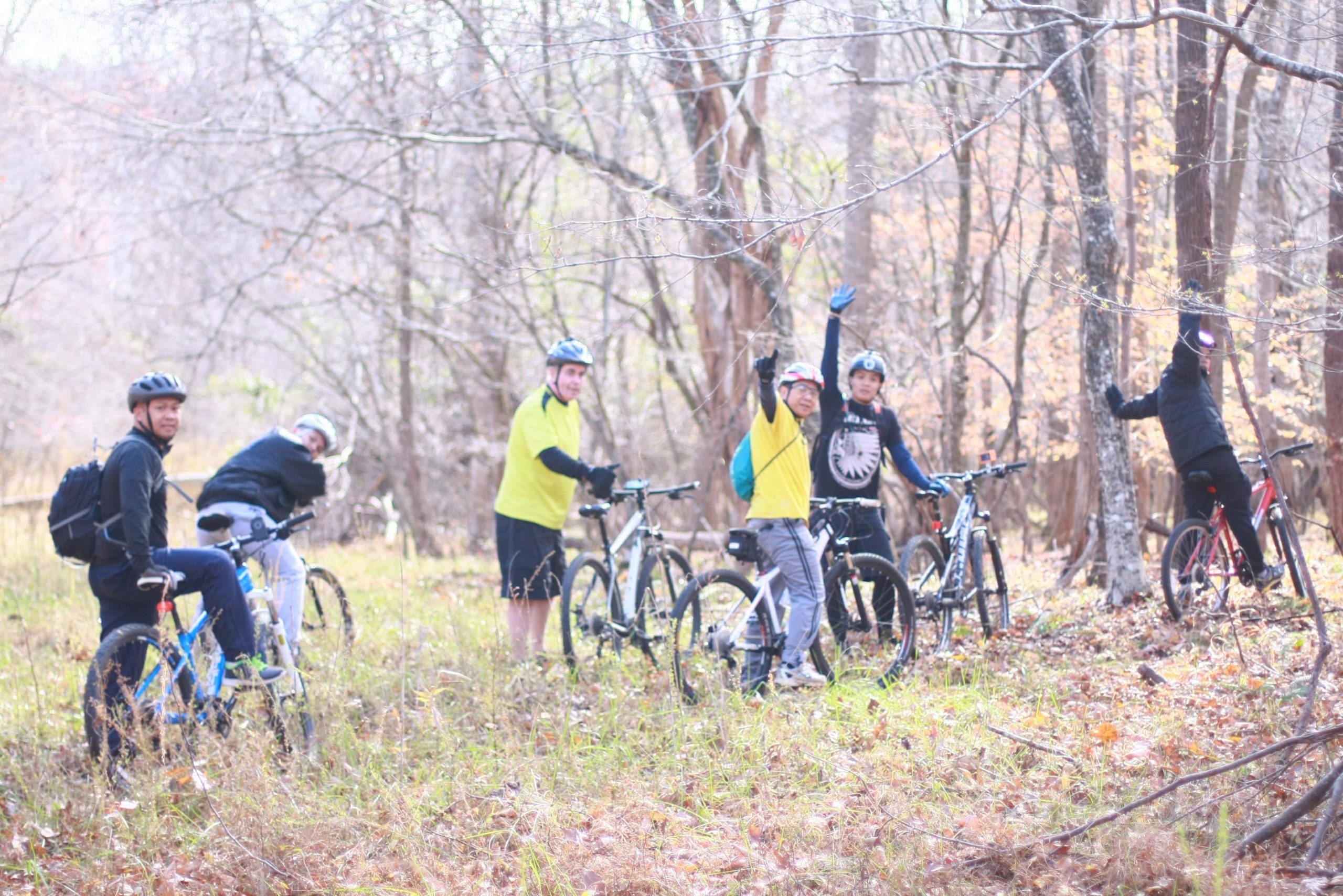 A group of six people wearing cycling helmets and colorful clothing, standing with bicycles in a forested area during autumn. Some individuals are smiling and waving, while others are seated or leaning against their bikes. The background features bare trees and dry grass, creating a seasonal atmosphere. Cedar Ridge Chatmoss mountain bike trail.