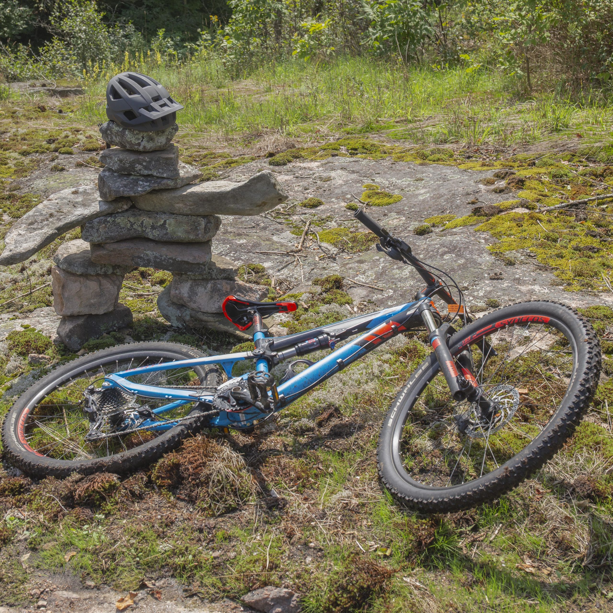 A mountain bike lies on its side on a rocky, moss-covered surface with a stack of stones beside it topped by a black helmet. The background features a green landscape with various plants and trees. All Trails mountain bike trail.