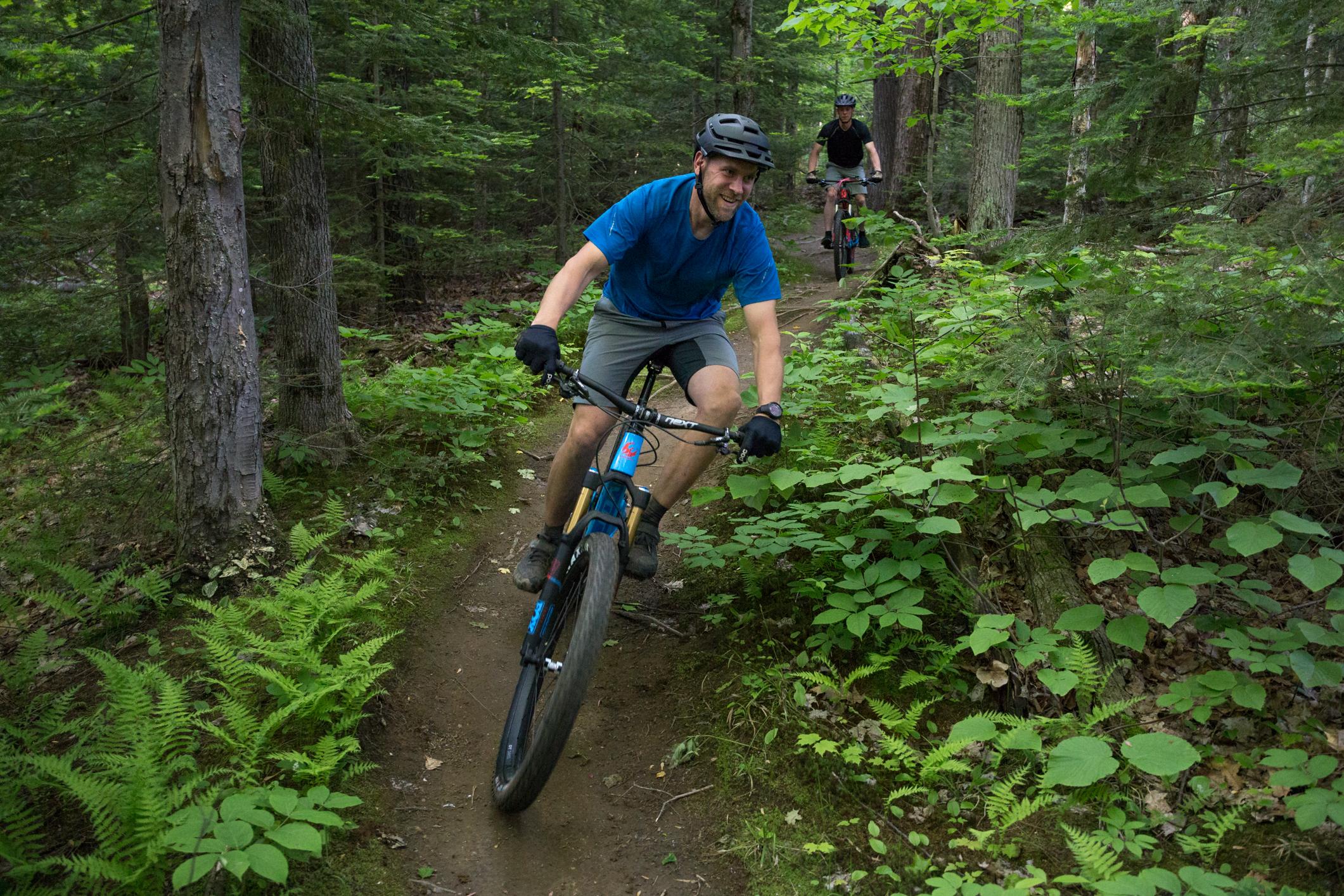 Pivot SwitchBlade: A cyclist in a blue shirt and gray shorts rides a mountain bike along a narrow trail surrounded by lush green foliage and trees in a forested area. Another cyclist in black can be seen riding in the background.