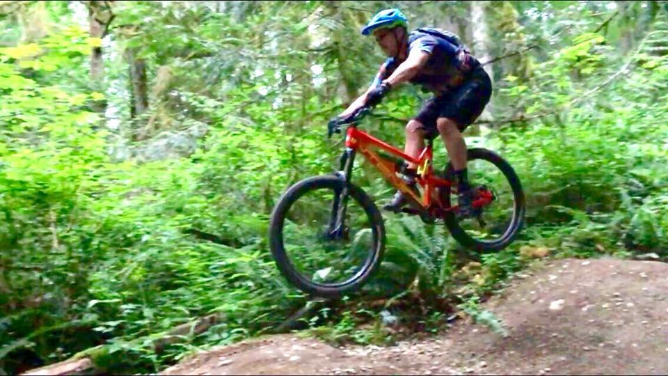 Intense Tracer 275c: A mountain biker dressed in a blue helmet and athletic wear jumps off a dirt ramp surrounded by lush green foliage in a forested area.