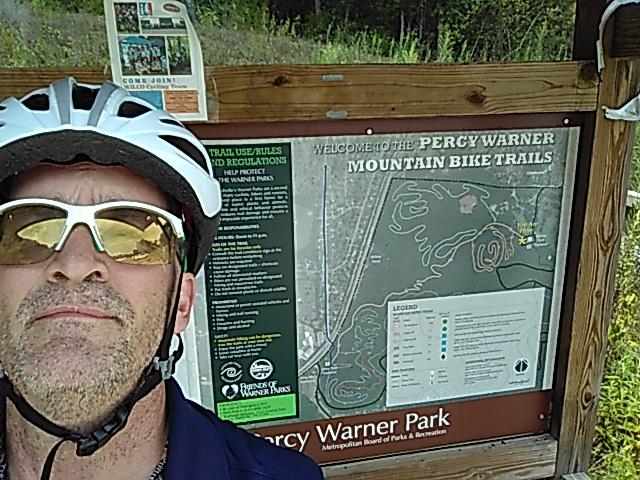 A person wearing a cycling helmet and sunglasses poses in front of a sign that reads "Welcome to the Percy Warner Mountain Bike Trails." The sign includes a map of the bike trails and information about park rules and regulations, with trees and greenery visible in the background. Percy Warner Mountain Bike Trails mountain bike trail.