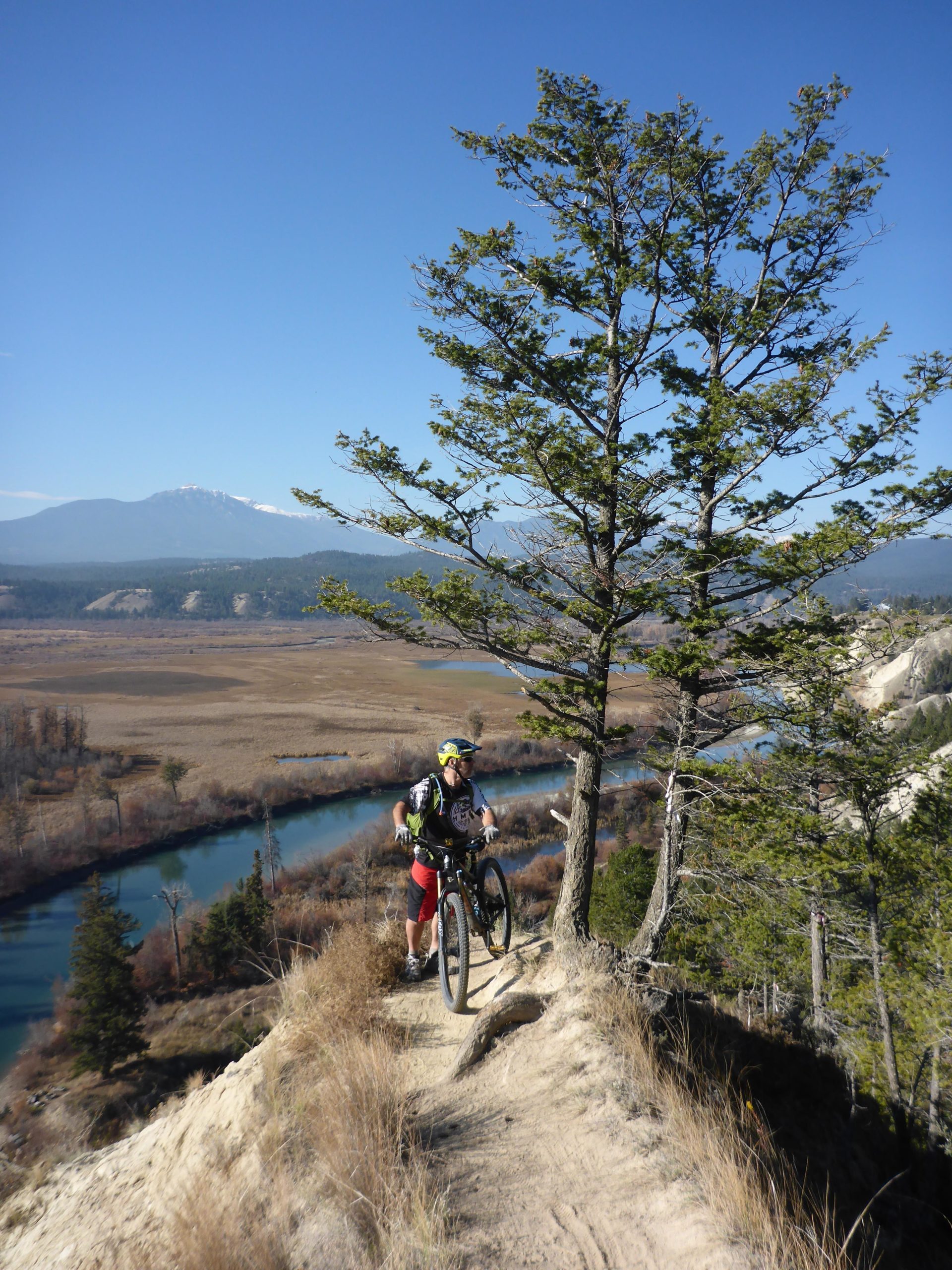 A mountain biker navigating a narrow trail along a hillside, with a scenic view of a river and valley below. The background features blue skies and distant mountains, while a tall tree provides shade on the right side of the image. Deja View mountain bike trail.