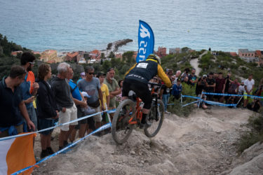 A mountain biker navigates a rocky downhill track while a large crowd watches from the sidelines. The backdrop features a coastal scene with colorful buildings along the shore and a calm sea. Blue and white flags are visible, indicating a competitive event. DH Uomini mountain bike trail.
