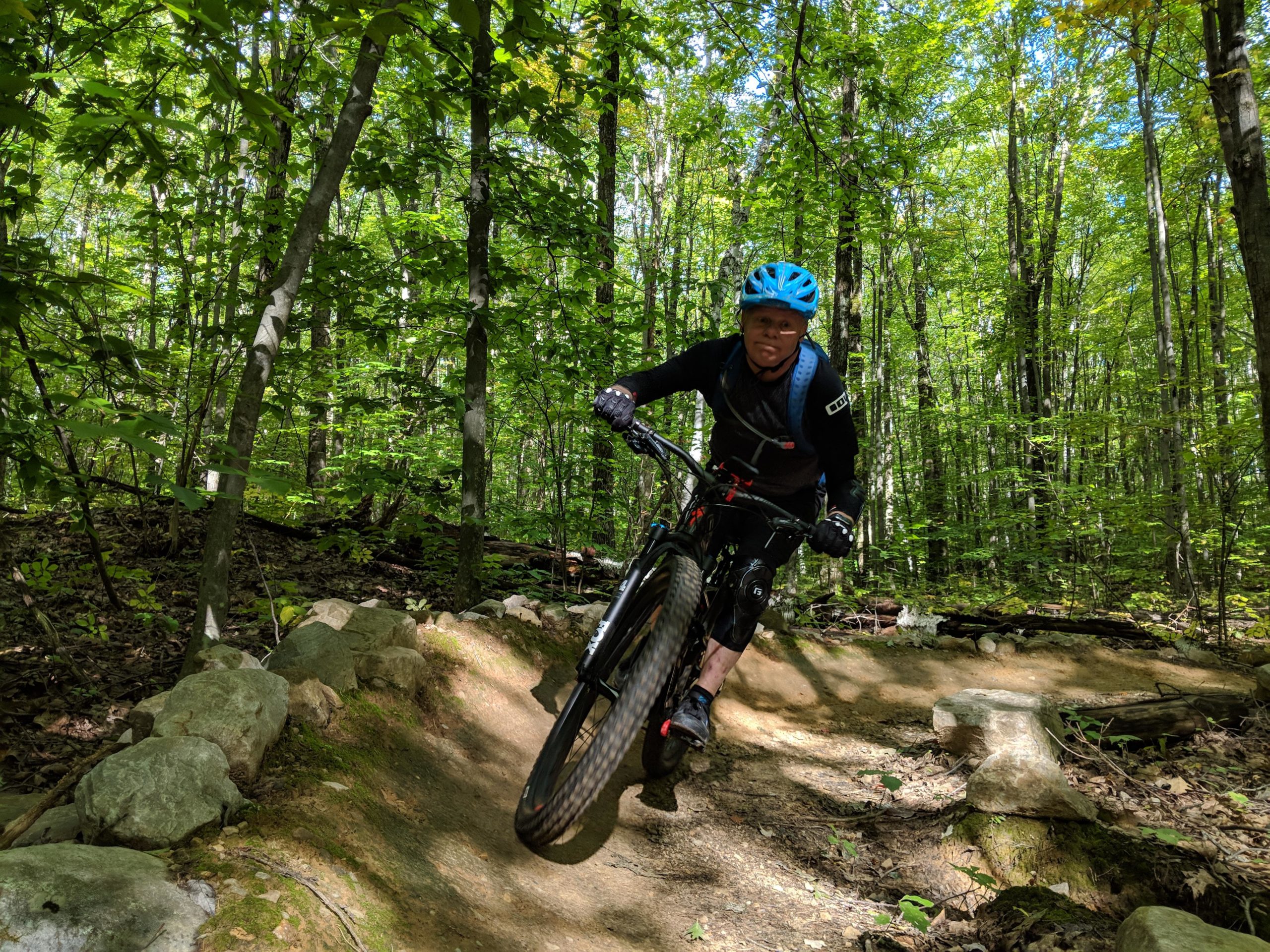 A mountain biker riding down a narrow dirt trail in a lush green forest, with tall trees surrounding the path. The biker is wearing a blue helmet and black cycling gear, leaning into a turn while maintaining speed. Sunlight filters through the leaves, illuminating the scene. Pine Hill Park mountain bike trail.
