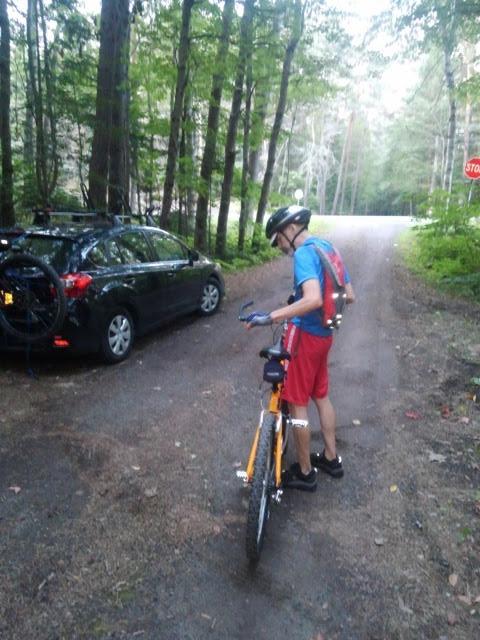 A person in a blue shirt and red shorts stands near a parked car on a gravel road, adjusting their orange bicycle. The setting is surrounded by tall trees, indicating a wooded area, with a stop sign visible in the distance. Slush Pond Trail mountain bike trail.