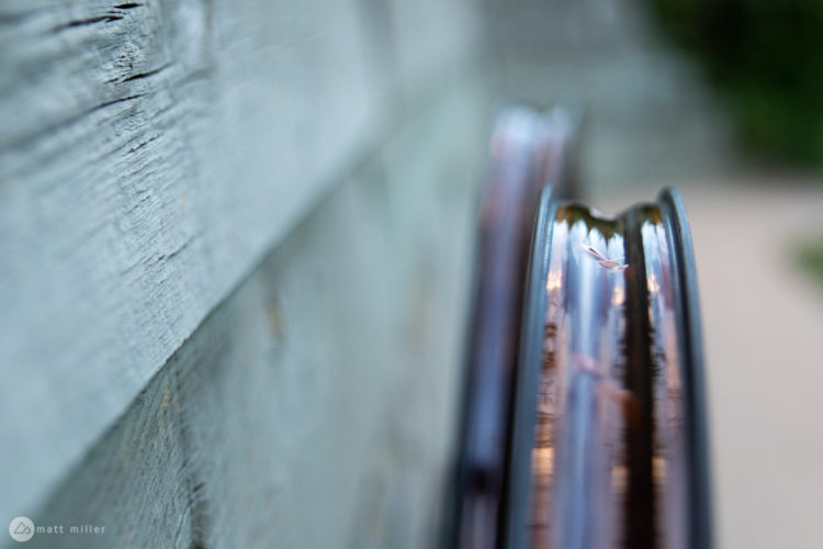 Close-up view of two car rims leaning against a textured wooden surface, with a soft focus on the rims reflecting ambient light. The wooden background features a weathered finish, highlighting its natural grain.