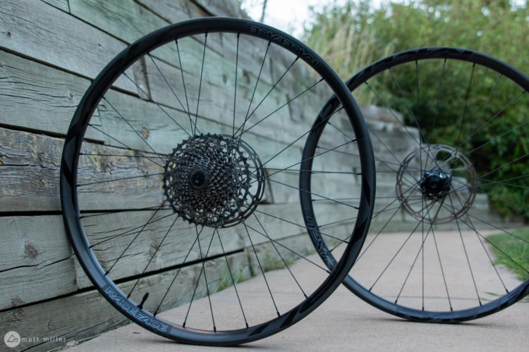 Two black bicycle wheels with disc brake rotors, positioned at an angle. The wheels are displayed against a weathered wooden background, showcasing their intricate spoke design and freehub. The image captures the essence of biking equipment, highlighting the wheels' sleek and modern aesthetic.