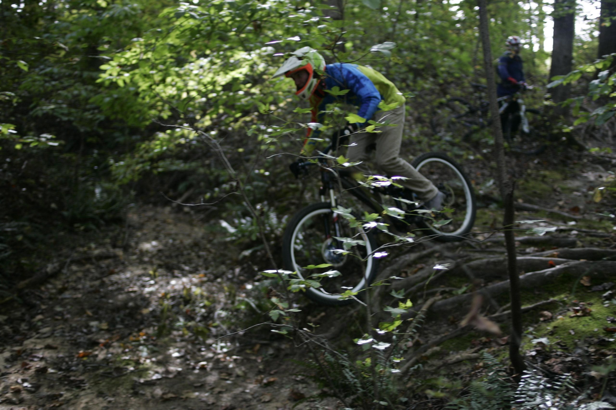 A mountain biker navigates a wooded trail, skillfully maneuvering over roots and uneven ground. Sunlight filters through the trees, casting dappled shadows on the path. Another cyclist can be seen in the background, waiting to continue. Deadfall Trail mountain bike trail.