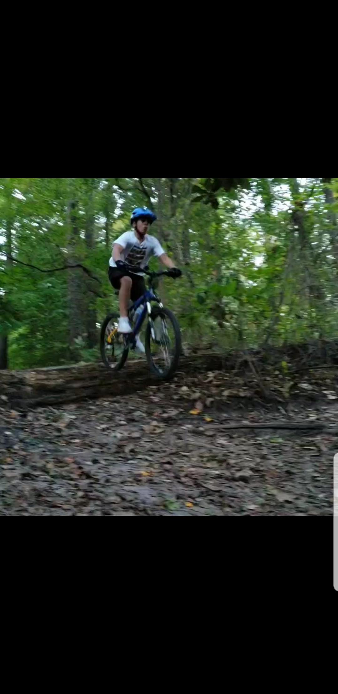 A person riding a mountain bike on a forest trail, with trees and greenery in the background. The rider is wearing a blue helmet and a t-shirt, navigating over a rough, leaf-covered path. Indian River Park mountain bike trail.
