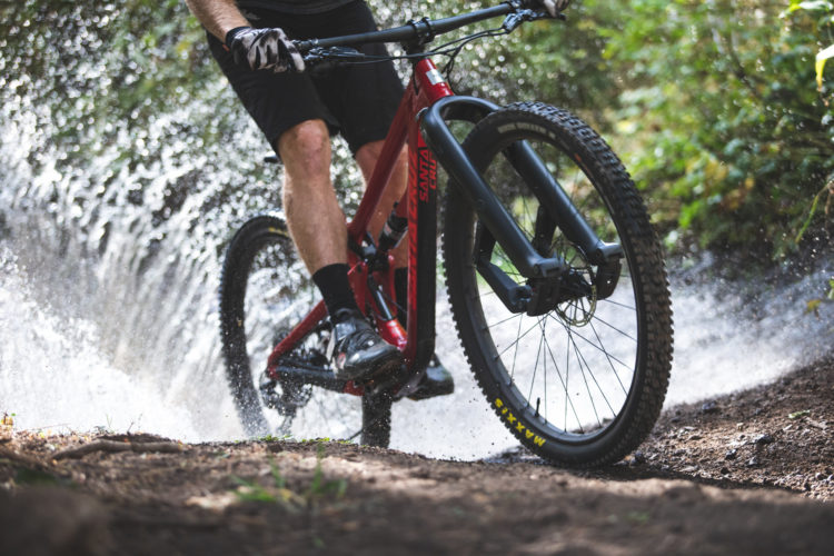 A close-up of a mountain biker navigating a muddy trail, splashing through water. The foreground features the bike's front wheel and the rider's legs, while droplets of water spray in the air, creating a dynamic and energetic scene. Lush greenery surrounds the path, emphasizing the outdoor environment.