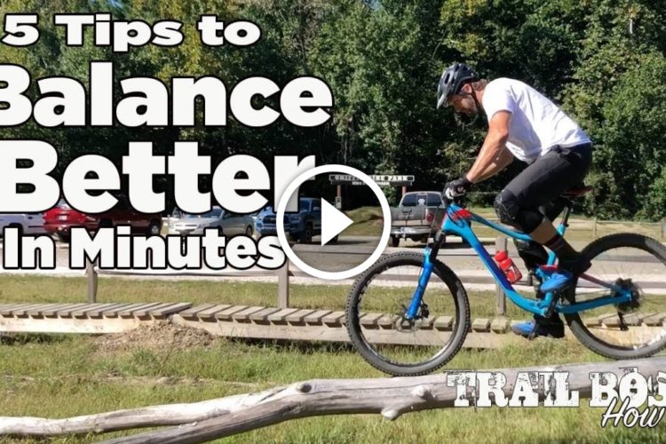 A person riding a mountain bike balances while maneuvering over a wooden beam in an outdoor setting, with vehicles parked in the background. The image features text overlay that reads "5 Tips to Balance Better In Minutes" along with a play button indicating a video tutorial.