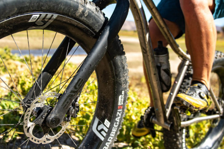 A close-up view of a mountain bike tire and frame, with a rider's leg visible. The bike features a wide, rugged tire suitable for off-road terrain and a disc brake system. Bright green foliage and a blurred landscape are in the background, indicating an outdoor setting.