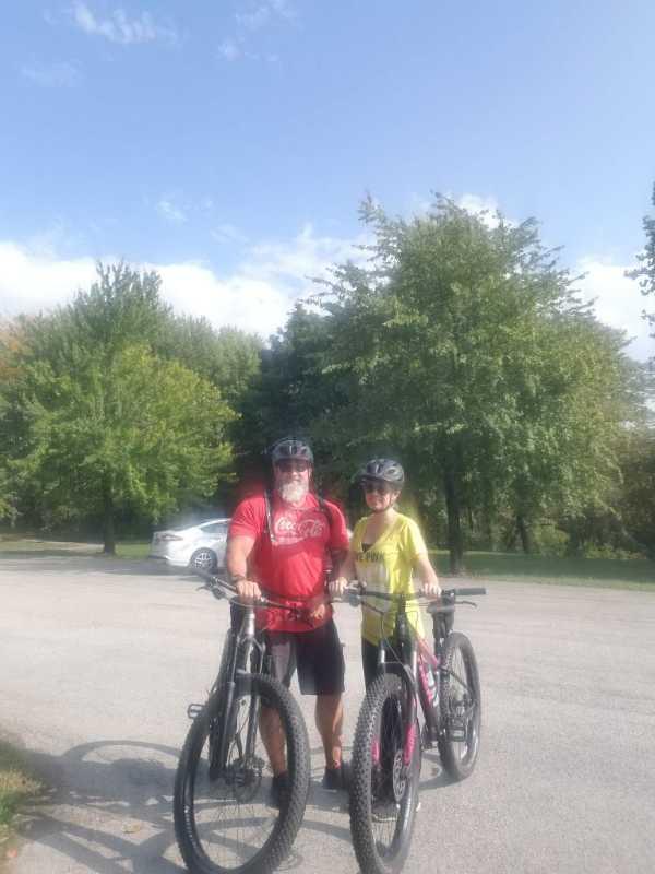 A man and a girl stand beside their mountain bikes in an outdoor setting, smiling at the camera. They are wearing helmets, with the man in a red shirt and the girl in a yellow shirt. Lush green trees are in the background under a clear blue sky. A parked car is visible in the distance. Alum Creek Phase II mountain bike trail.