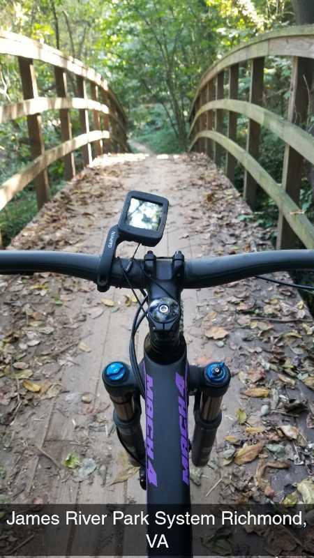 A view from the handlebars of a mountain bike on a wooden bridge surrounded by trees, with a clear path ahead. The bike's computer display is visible, showing data. Fallen leaves cover the ground, indicating a scenic ride through the James River Park System in Richmond, VA. Buttermilk mountain bike trail.