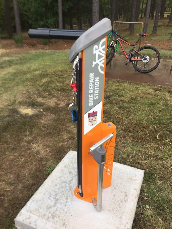 A bike repair station with various tools attached, including a tire pump and repair tools, set on a concrete base surrounded by grass and trees. In the background, a bicycle is visible. The station has a sign that reads "BIKE REPAIR STATION." Cedar Glades Trail mountain bike trail.