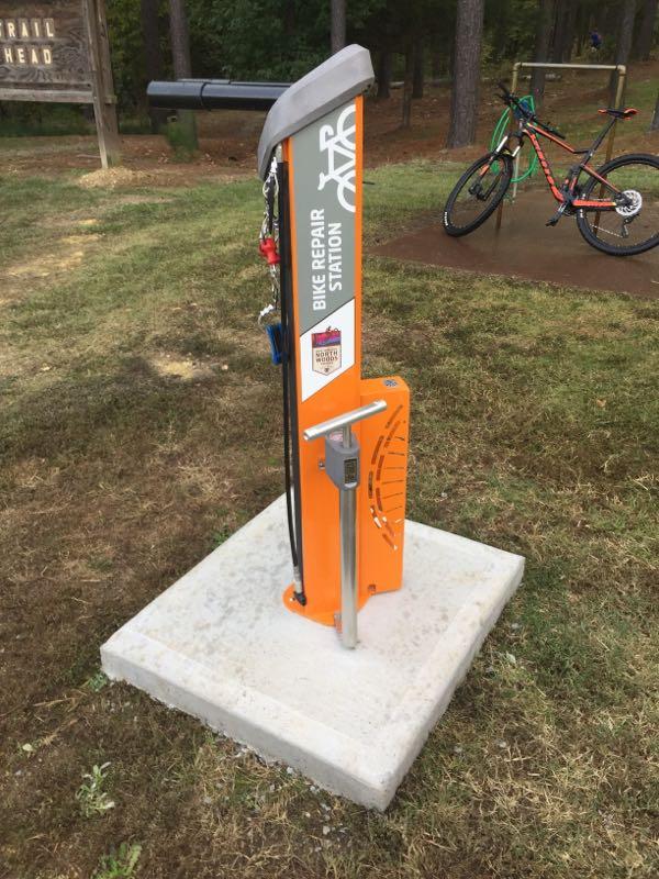 An orange bike repair station with a pump and tools attached, set on a concrete base in a grassy area. In the background, a sign indicates a trailhead, and a colorful bicycle is parked nearby. Cedar Glades Trail mountain bike trail.