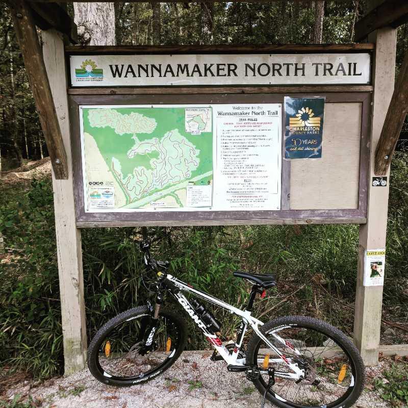 A sign for the Wannamaker North Trail featuring a detailed map of the trail system. A mountain bike is parked in front, with lush greenery surrounding the area. The sign includes trail rules and information about the park's history. Wannamaker Park mountain bike trail.