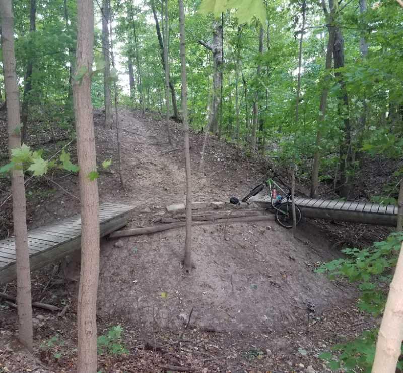 A forest scene featuring a dirt path leading up a hill, flanked by trees. In the foreground, there is a wooden bridge crossing over a small section of the path. A bicycle is resting on the ground nearby, surrounded by sparse underbrush and foliage. The setting is lush with green leaves, indicating a healthy forest environment. Van  Buren mountain bike trail.