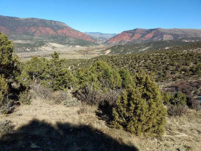 A panoramic view of a mountainous landscape featuring rolling hills and vibrant red rock formations. In the foreground, there are patches of greenery including shrubs and trees, while the background showcases a clear blue sky and distant mountain ranges. The scene captures the natural beauty and rugged terrain of the area. The Boneyard mountain bike trail.