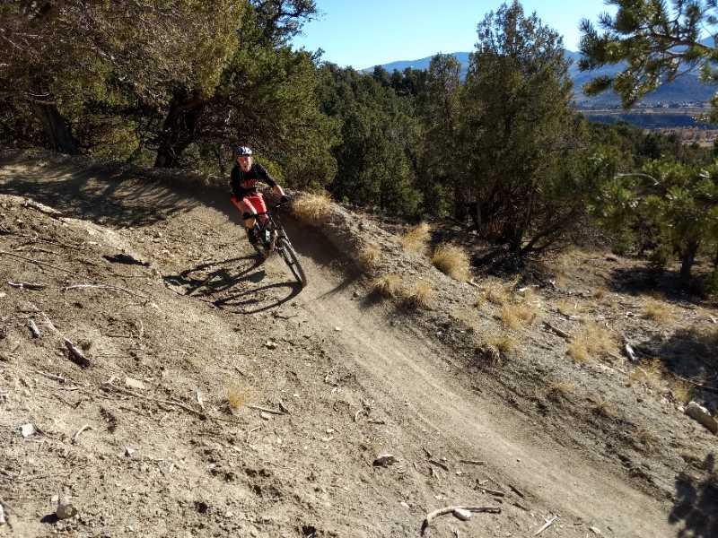 A mountain biker navigating a dirt trail on a hillside, surrounded by trees and mountains in the background. The biker is leaning into a turn, showcasing an action-oriented scene in a natural outdoor setting. Pool/ice Rink mountain bike trail.