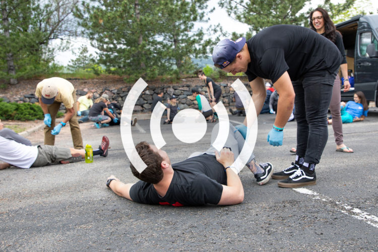An outdoor training session where individuals are practicing first aid techniques. Some participants are lying on the ground, while others are administering care or assisting them. The scene includes a mix of people, wearing casual clothing and gloves, engaged in various activities focused on emergency response skills. Trees and a vehicle are visible in the background, suggesting a community or outdoor setting.