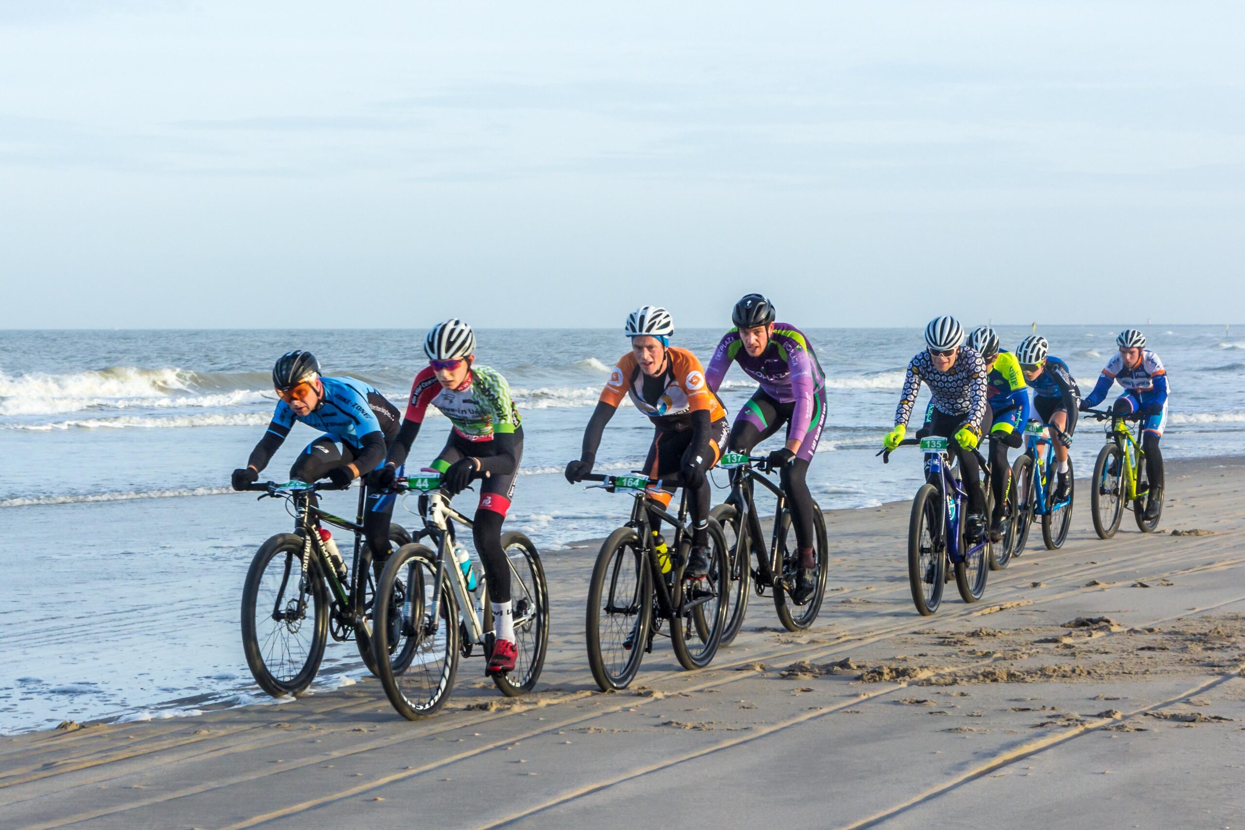 A group of six cyclists riding beach bikes along a sandy shoreline, with ocean waves in the background. The cyclists are wearing colorful jerseys and helmets, showcasing a mix of riding styles and techniques as they navigate the terrain.