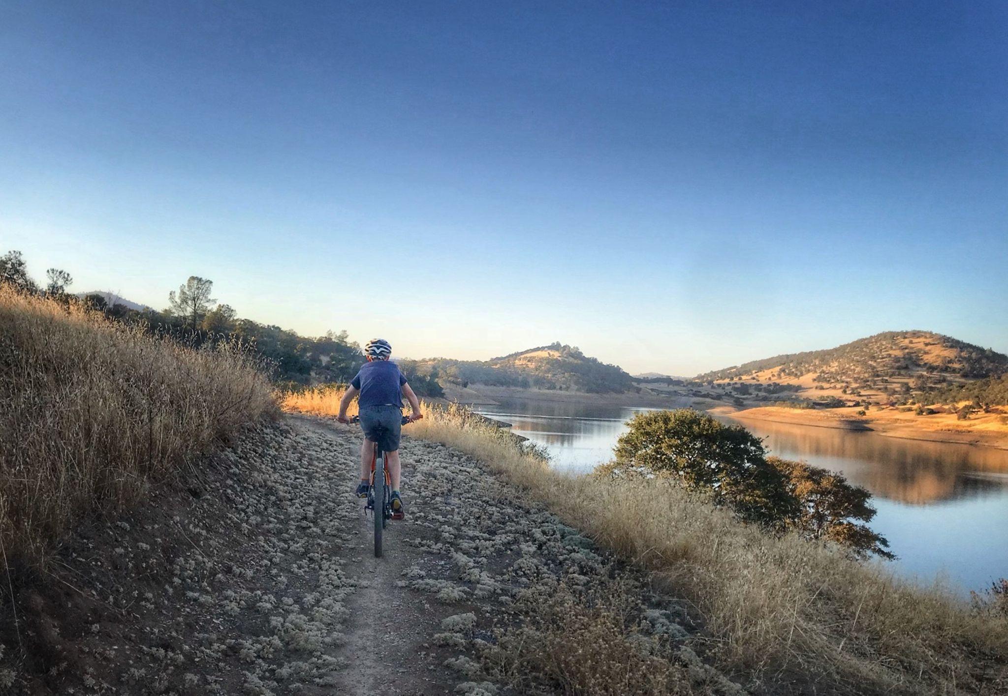 A child riding a bicycle on a dirt path beside a tranquil lake, surrounded by tall grass and rolling hills in the background. The sky is clear with a hint of blue, reflecting the peacefulness of the outdoor scenery. Glory Hole mountain bike trail.