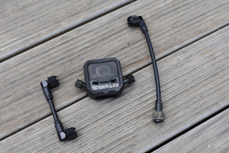 A black ShockWiz device with two connecting hoses lies on a wooden surface. The device features a square design with a prominent "Q" logo and the name "ShockWiz" displayed on it. One hose is curved with a threaded end, while the other is straight with fittings on both ends.
