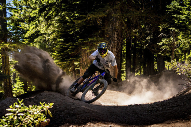 A mountain biker navigates a curved dirt trail in a forested area, kicking up a cloud of dust. The rider wears a helmet and protective gear, and is captured in action as they lean into the turn, surrounded by tall trees and sunlight filtering through the leaves.