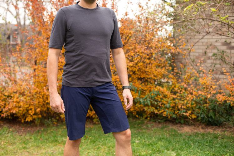 A person standing outdoors wearing a dark gray short-sleeve athletic shirt and navy blue shorts, with a watch on their left wrist. In the background, there is a colorful foliage of orange and yellow leaves, indicating a fall season.