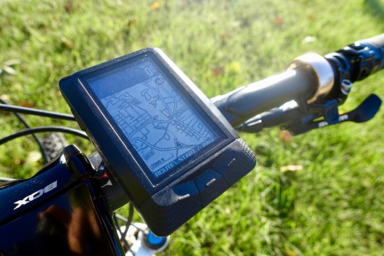A close-up view of a bike's handlebar with a GPS device displaying a map. The screen shows a detailed route overlay with an elevation of 12,000 feet indicated. The background features green grass, suggesting an outdoor setting.