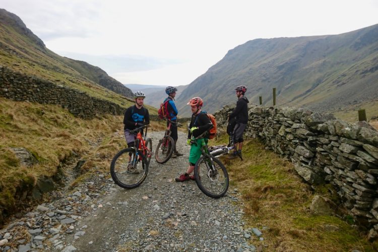 Four mountain bikers are paused on a rocky path in a scenic valley, surrounded by green hills and stone walls. Two riders are standing next to their bikes, while another leans against a stone wall. The group appears to be engaged in conversation, with a cloudy sky overhead. The landscape is rugged and natural, showcasing the beauty of outdoor sports.