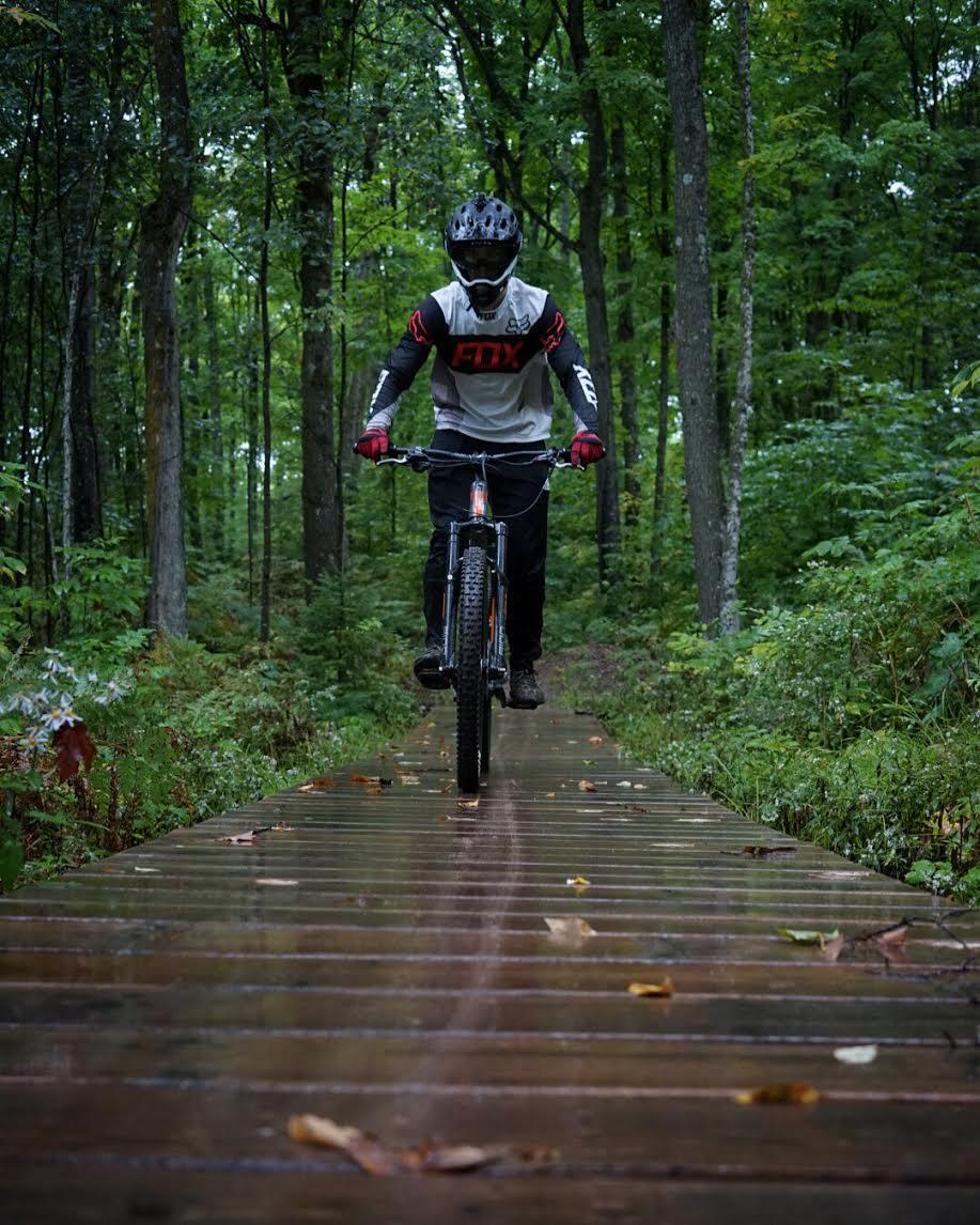 A person wearing a black helmet and protective gear rides a mountain bike on a wooden path surrounded by lush green trees and foliage. The trail is wet, likely from recent rain, with fallen leaves scattered across the wooden planks. WinMan Trail mountain bike trail.