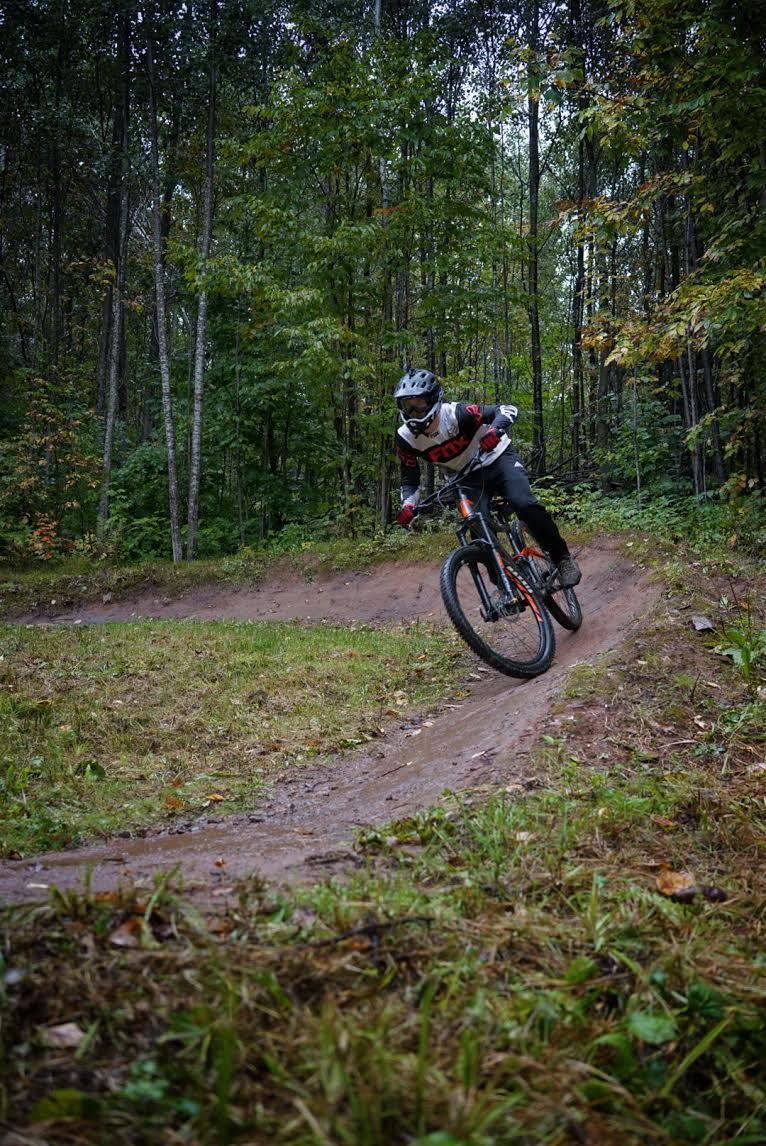 A mountain biker performing a sharp turn on a dirt trail surrounded by lush green trees and underbrush, capturing the action and excitement of outdoor cycling. WinMan Trail mountain bike trail.