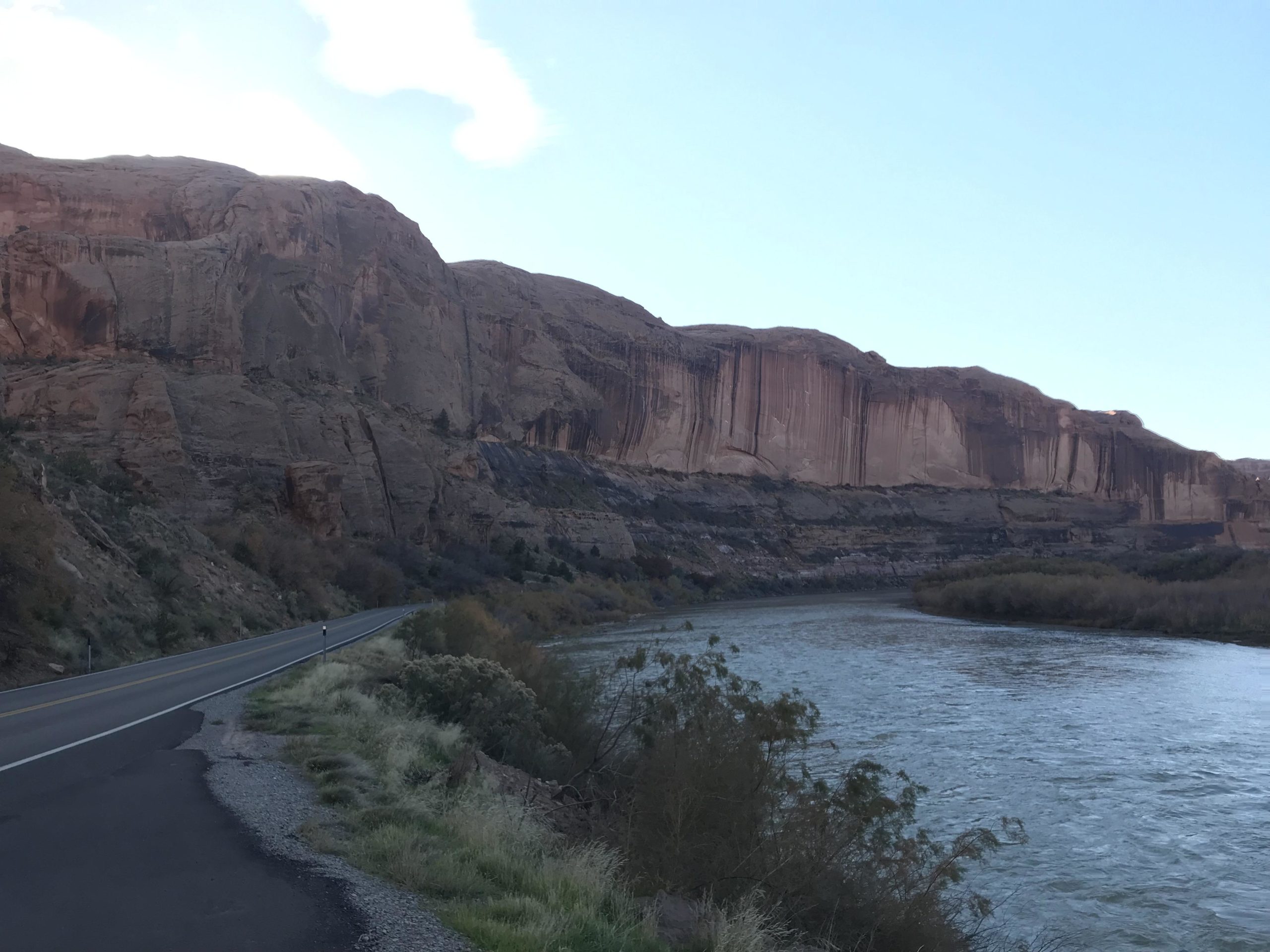A scenic view of a river flowing alongside a road, with towering rock formations and cliffs bordered by lush vegetation. The sky is partly cloudy, creating a dramatic backdrop to the natural landscape. Porcupine Rim mountain bike trail.