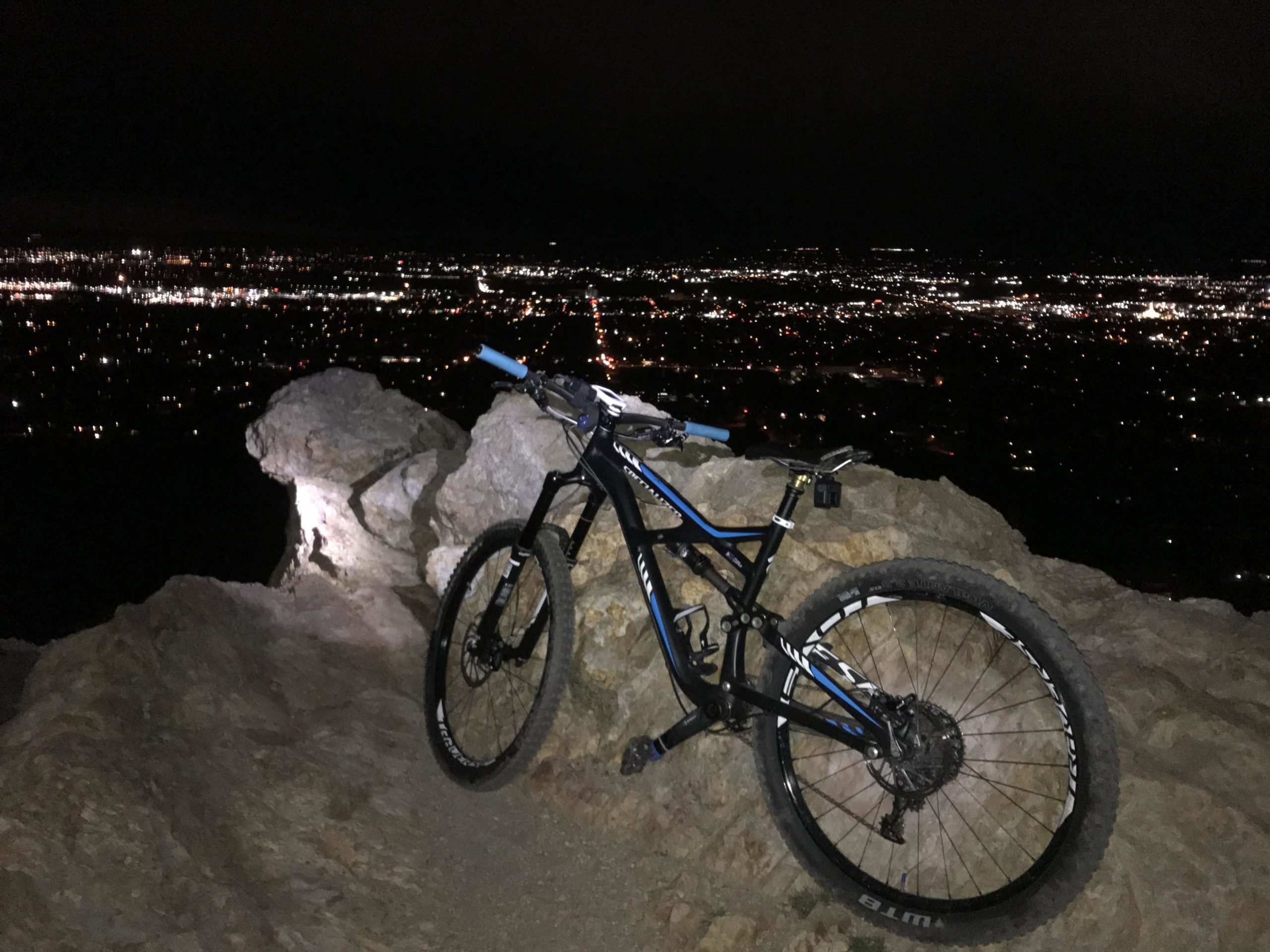 A mountain bike parked on a rocky outcrop at night, overlooking a city illuminated by countless lights in the distance. The scene captures the contrast between the rugged terrain and the vibrant urban landscape below. Bonneville Shoreline Trail - Ogden Section mountain bike trail.