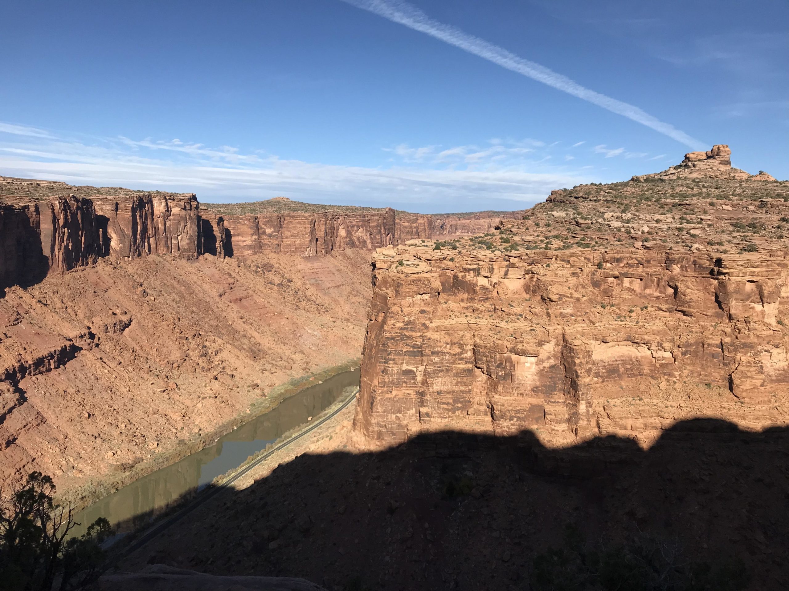 A sweeping view of a rocky canyon landscape featuring steep cliffs and a winding river at the base. The sky is clear with wispy clouds, and the sun casts shadows across the rugged terrain. Porcupine Rim mountain bike trail.
