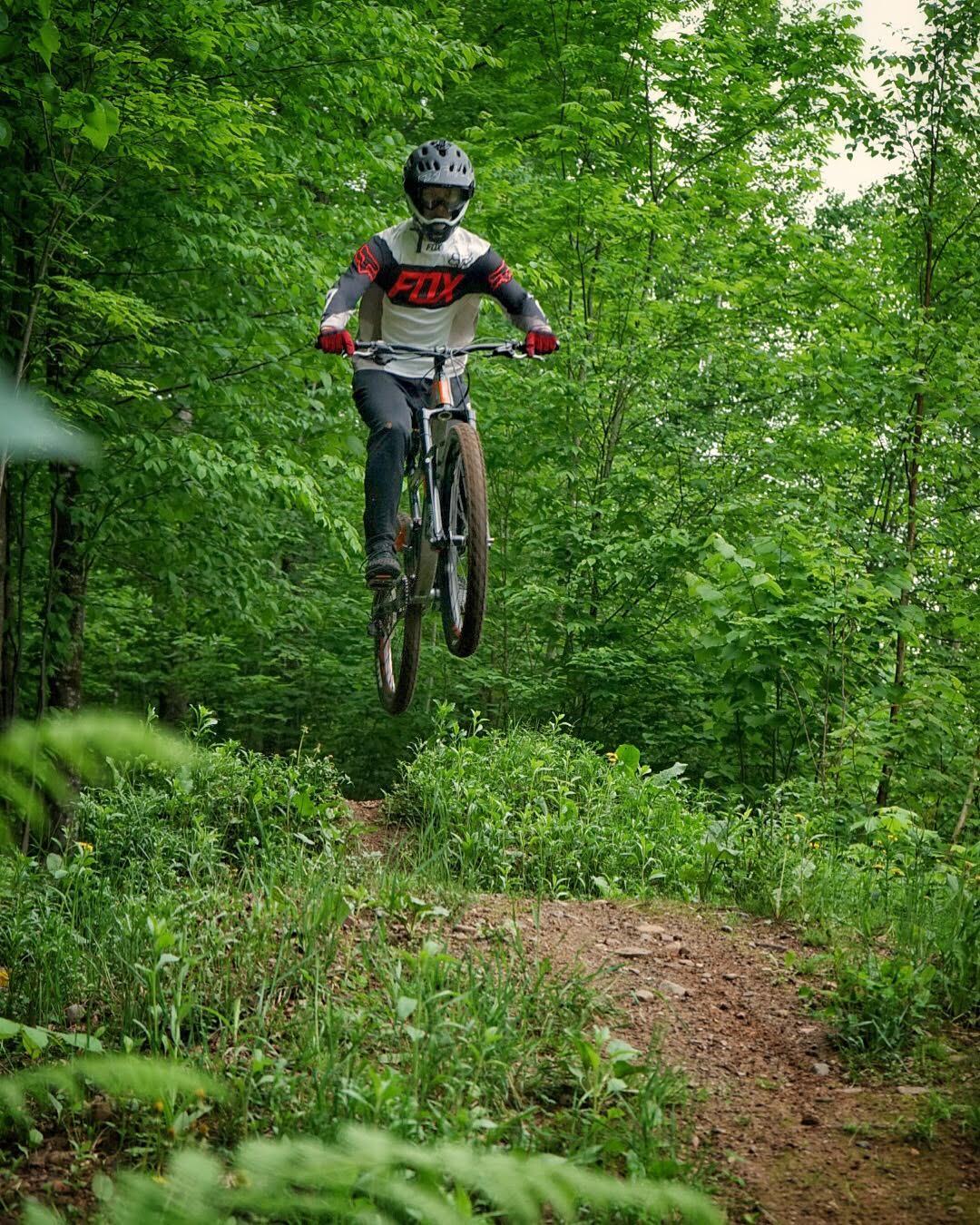 A mountain biker in a helmet and protective gear performs a jump on a dirt trail surrounded by lush greenery. The rider is mid-air, showcasing an energetic moment in mountain biking. WinMan Trails mountain bike trail.