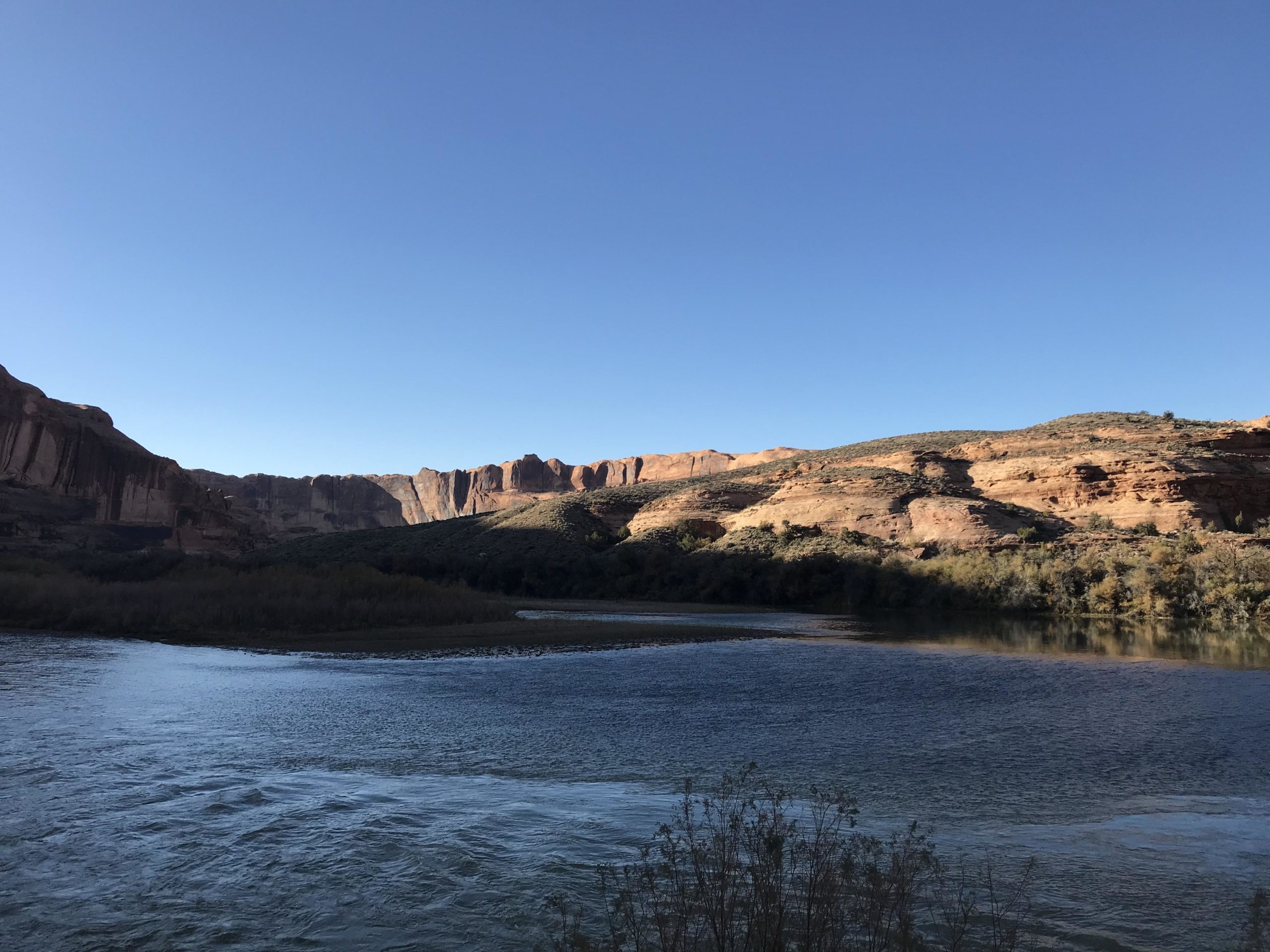 A serene river flowing through a canyon landscape, surrounded by rugged rock formations and green vegetation under a clear blue sky. The water reflects the sunlight, creating a peaceful atmosphere. Porcupine Rim mountain bike trail.